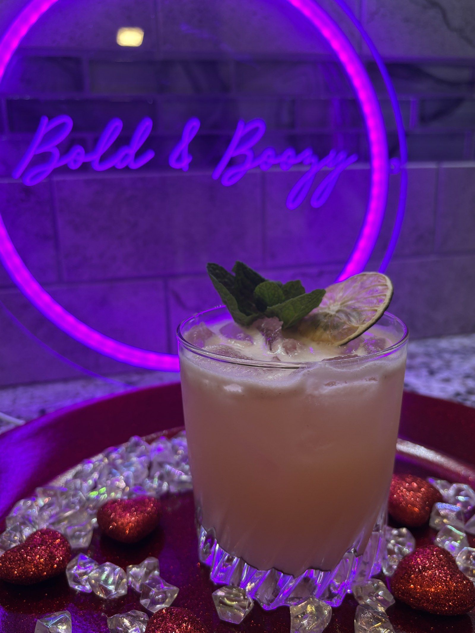 A creamy pink drink with mint and a dried lime wheel in front of the glowing Bold & Boozy neon sign