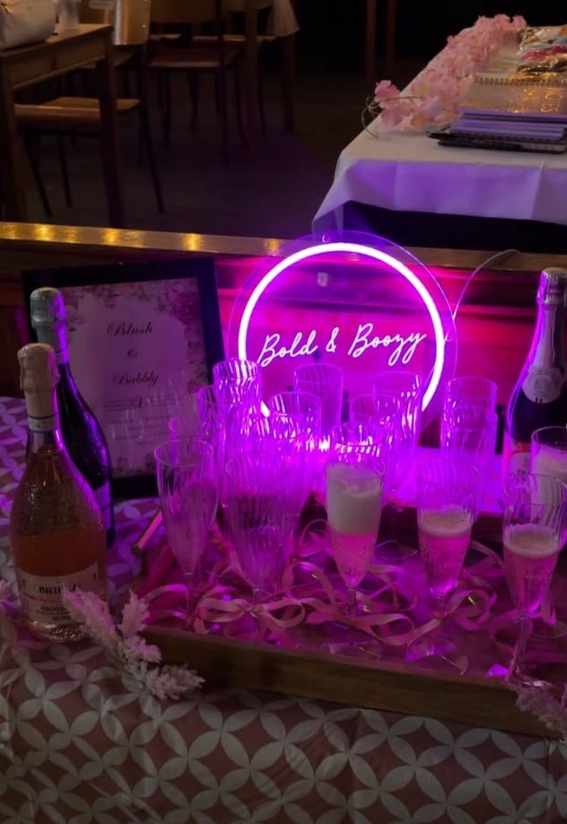 A sparkling wine station with a glowing Bold & Boozy neon sign, ribbons and a tray of flutes