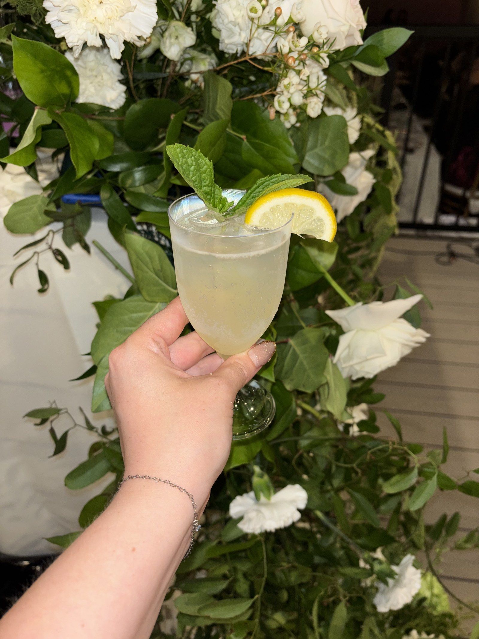 A pale cocktail with a lemon wheel and mint, held up in front of white flowers